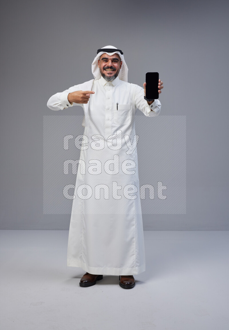 Saudi man Wearing Thob and white Shomag standing showing phone to camera on Gray background