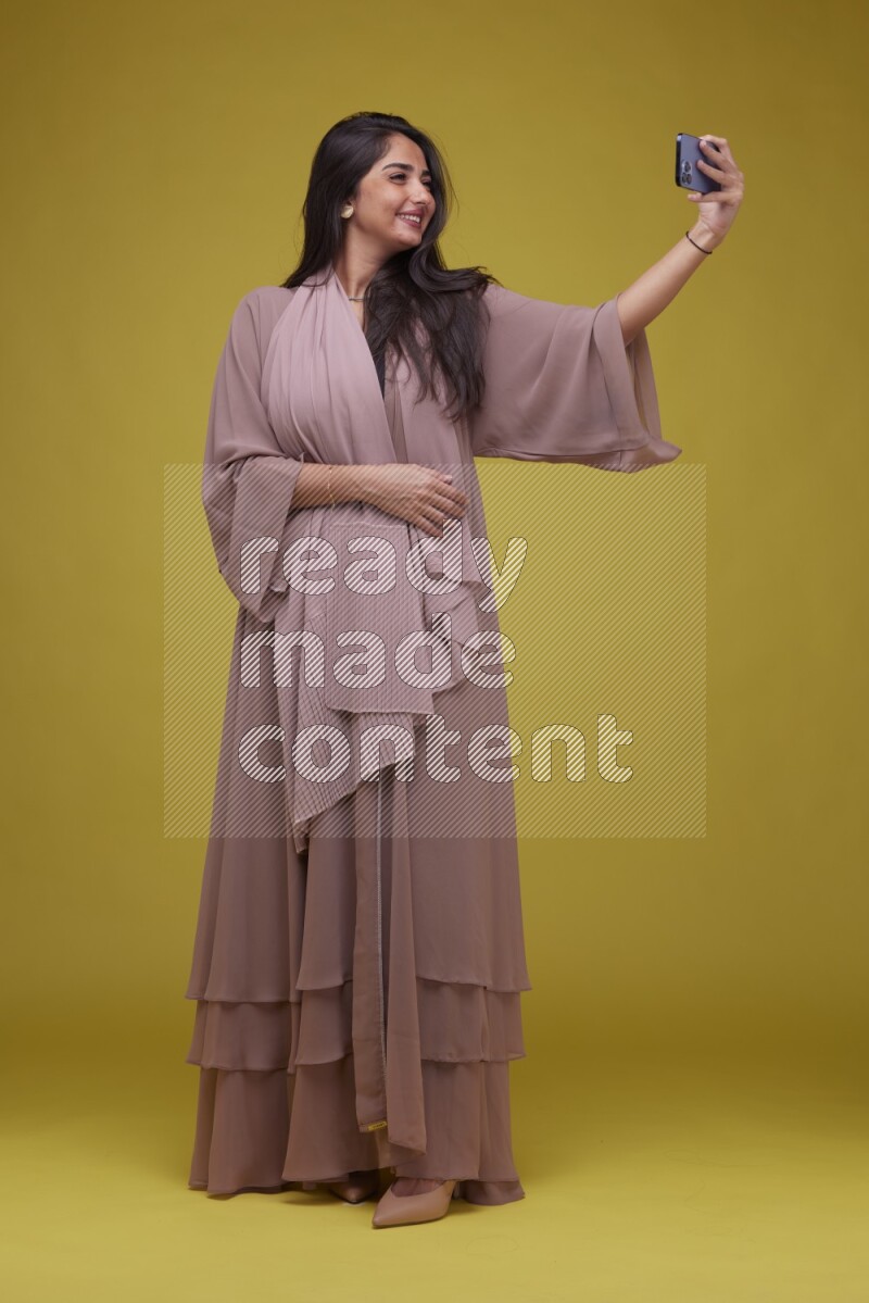 A woman Taking a Selfie on a Yellow Background wearing Brown Abaya