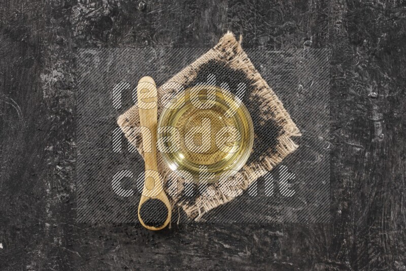 A glass bowl full of black seeds oil and wooden spoon full of black seeds with seeds spreaded on burlap fabric on a textured black flooring