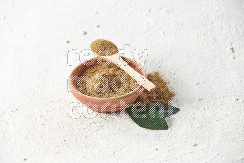 A wooden bowl and wooden spoon full of cumin powder on textured white flooring