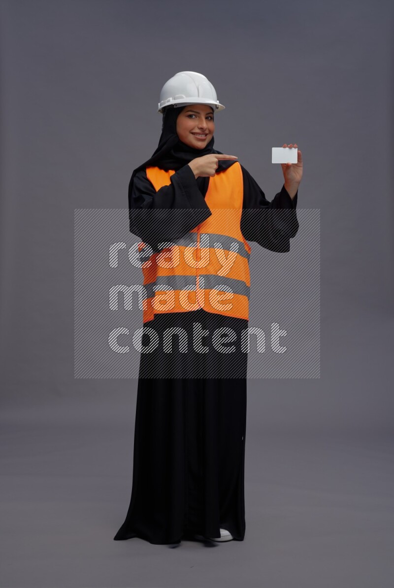 Saudi woman wearing Abaya with engineer vest standing holding ATM card on gray background