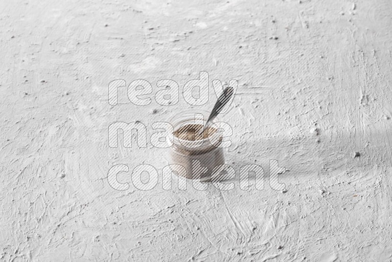 A glass jar full of black pepper powder and a metal spoon full of powder on a textured white flooring