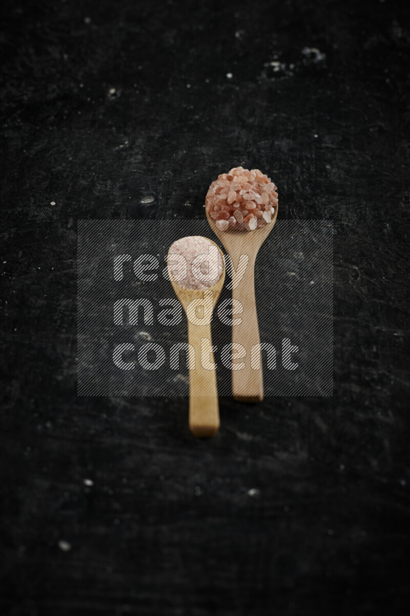 2 wooden spoons filled with fine and coarse salt on black background