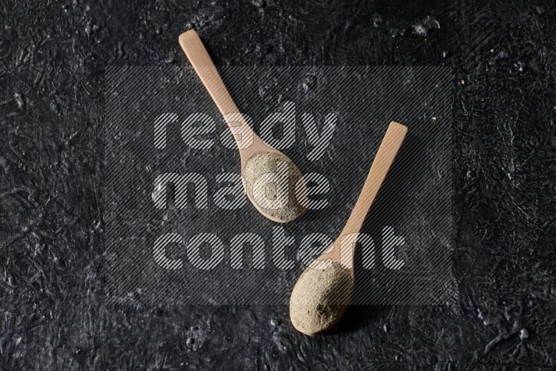 2 wooden spoons full of white pepper powder on textured black flooring