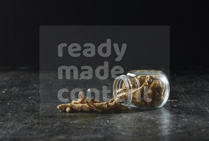 A flipped glass spice jar full of dried turmeric whole fingers and fingers fell out of it on textured black flooring