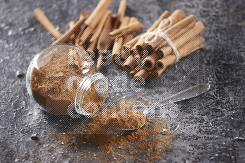 Herbal glass jar full cinnamon powder flipped and a metal spoon full of powder, cinnamon sticks stacked and bounded in the back on textured black background in different angles