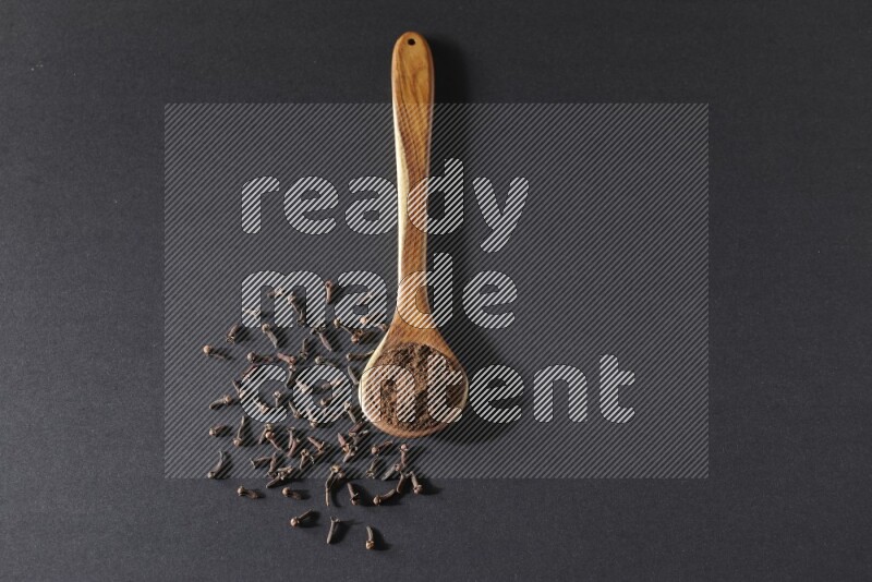 A wooden ladle full of cloves powder and some of whole cloves around it on a black flooring
