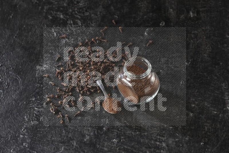 A glass spice jar and a metal spoon full of cloves powder with cloves spread on textured black flooring