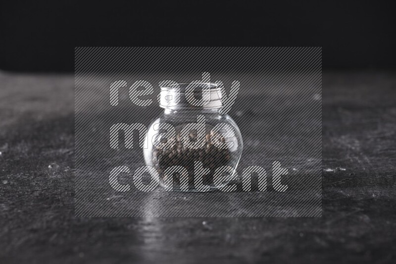 A glass spice jar full of black pepper beads on textured black flooring