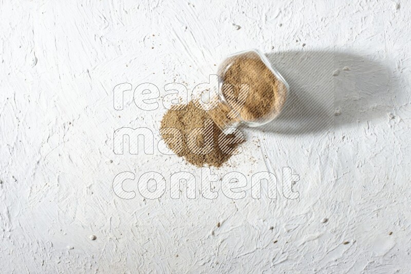 وعاء زجاجي مقلوب ممتلئ ببودرة الكمون والبودرة تناثرت منه على خلفية بيضاء