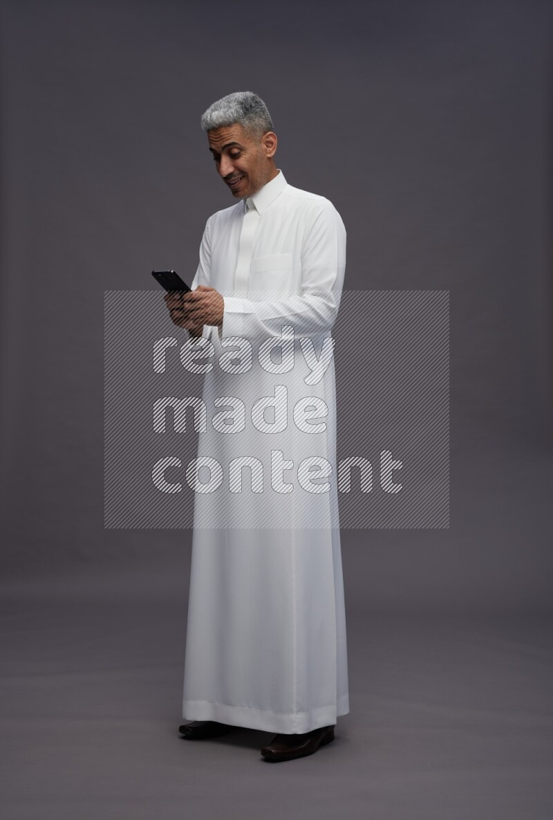 Saudi man wearing thob standing texting on phone on gray background