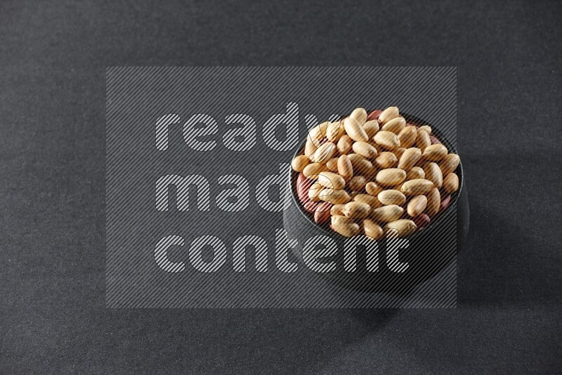 A black pottery bowl full of peeled peanuts on a black background in different angles