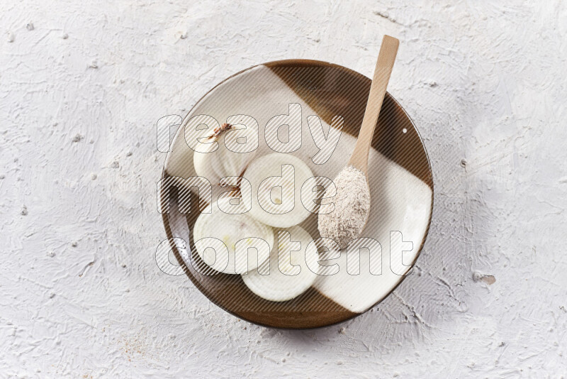 A wooden spoon full of onion powder with sliced onions, all on a pottery plate on white background