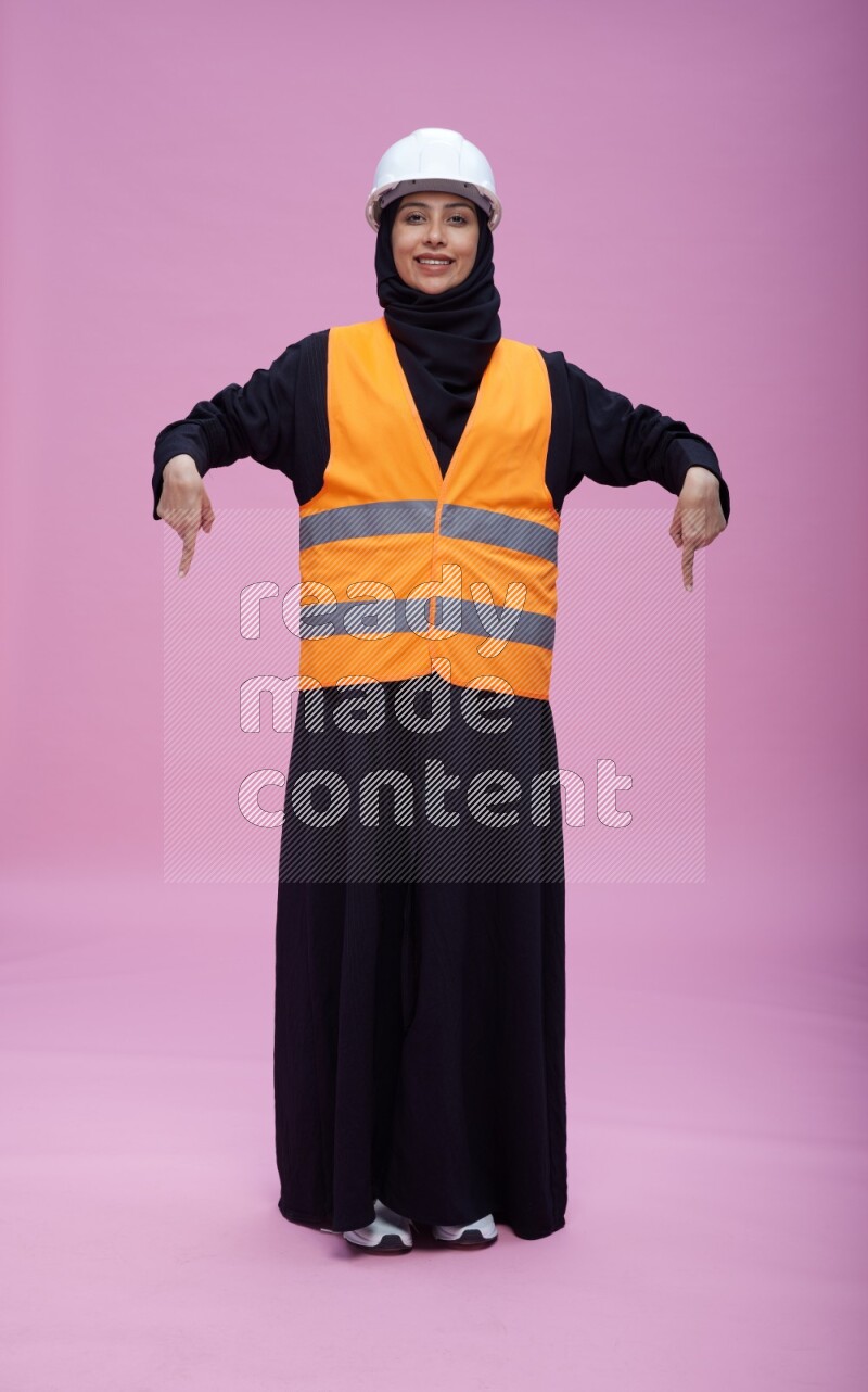 Saudi woman wearing Abaya with engineer vest and helmet standing interacting with the camera on pink background