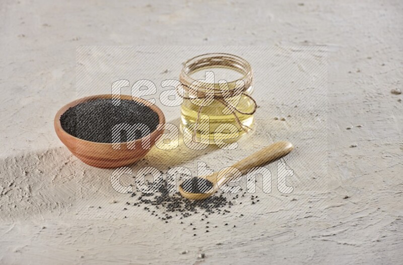 A wooden bowl and spoon full of black seeds with a glass jar of black seeds oil on a textured white flooring