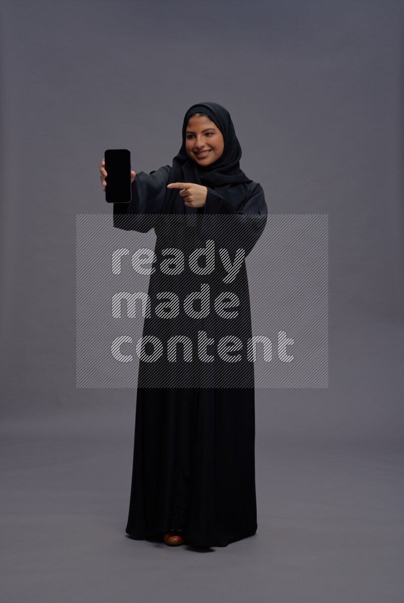 Saudi woman wearing Abaya standing showing phone to camera on gray background