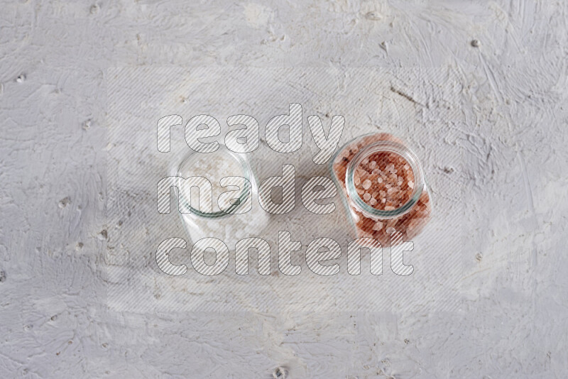 2 glass jars one is filled with coarse himalayan salt and the other with coarse sea salt on white background