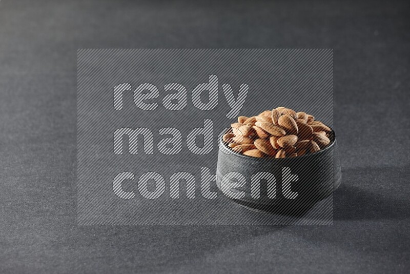 A black pottery bowl full of peeled almonds on a black background in different angles