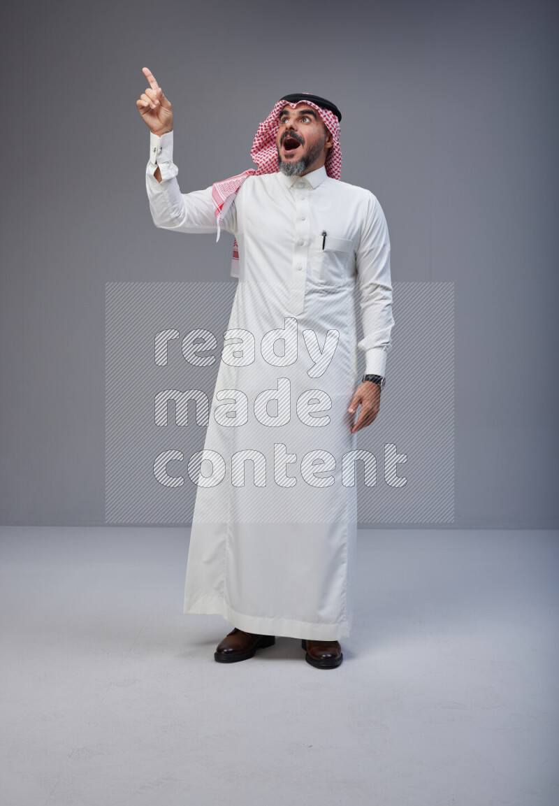 Saudi man Wearing Thob and red white Shomag standing interacting with the camera on Gray background