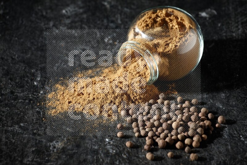 وعاء زجاجي للتوابل مقلوب ممتلئ ببودرة بهار الفلفل الحلو والبودرة تناثرت منه مع الحبوب على خلفية سوداء