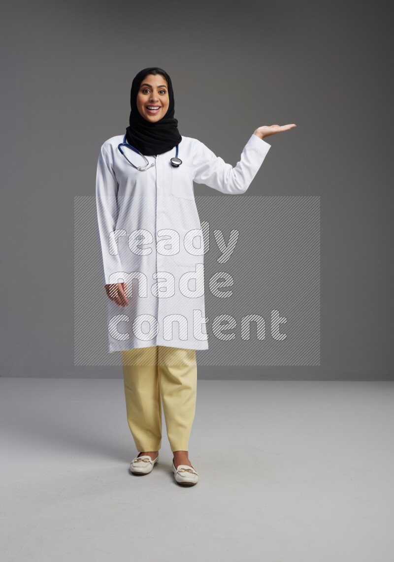 Saudi woman wearing lab coat with stethoscope standing interacting with the camera on Gray background
