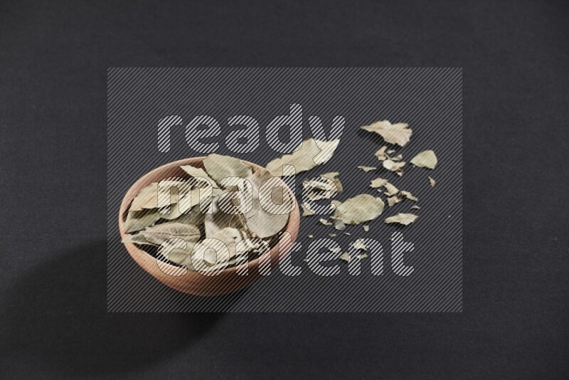 A wooden bowl filled with dried bay leaves on black flooring