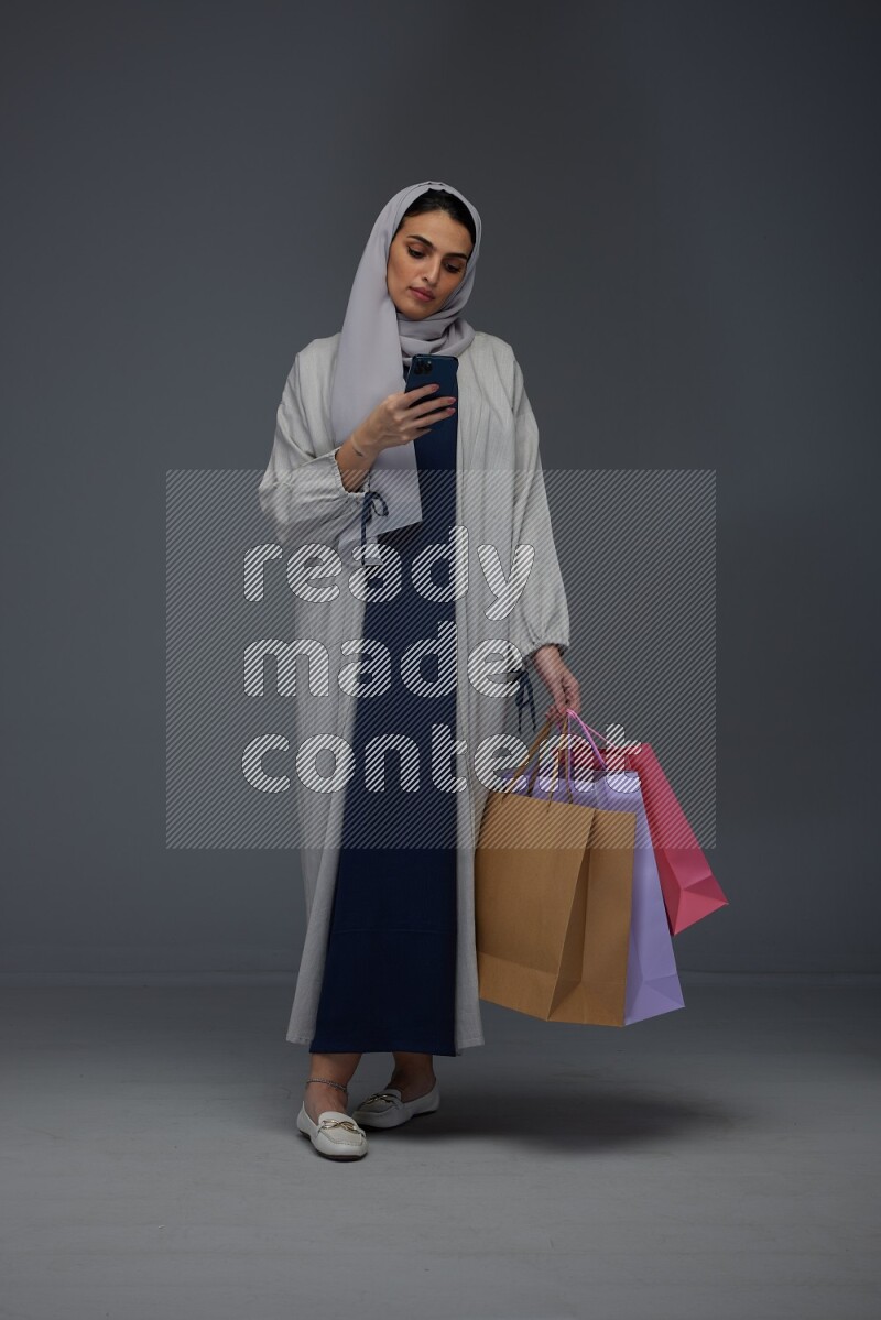 A Saudi woman wearing a light gray Abaya and head scarf standing and holding shopping bags making different poses eye level on a grey background