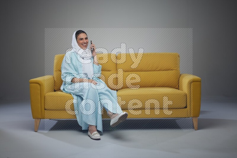 A Saudi woman wearing a light blue Abaya and a white head scarf sitting on a yellow sofa and talking in the phone while making different poses eye level on a grey background