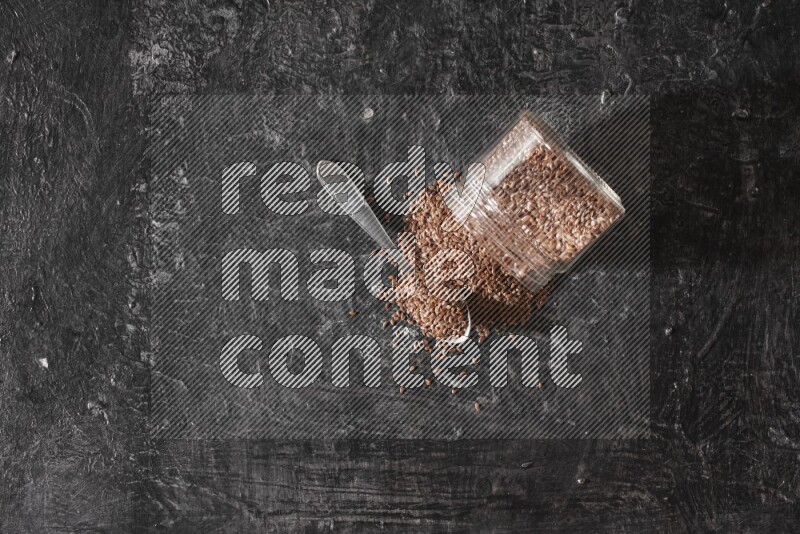 A glass jar full of flaxseeds flipped and seeds spread out with a metal spoon full of the seeds on a textured black flooring