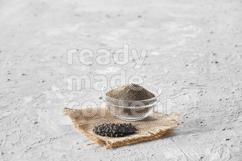 A glass bowl full of black pepper powder and black pepper beads on burlap fabric on textured white flooring