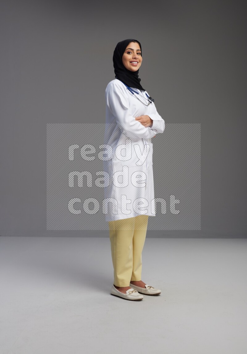 Saudi woman wearing lab coat with stethoscope standing with crossed arms on Gray background
