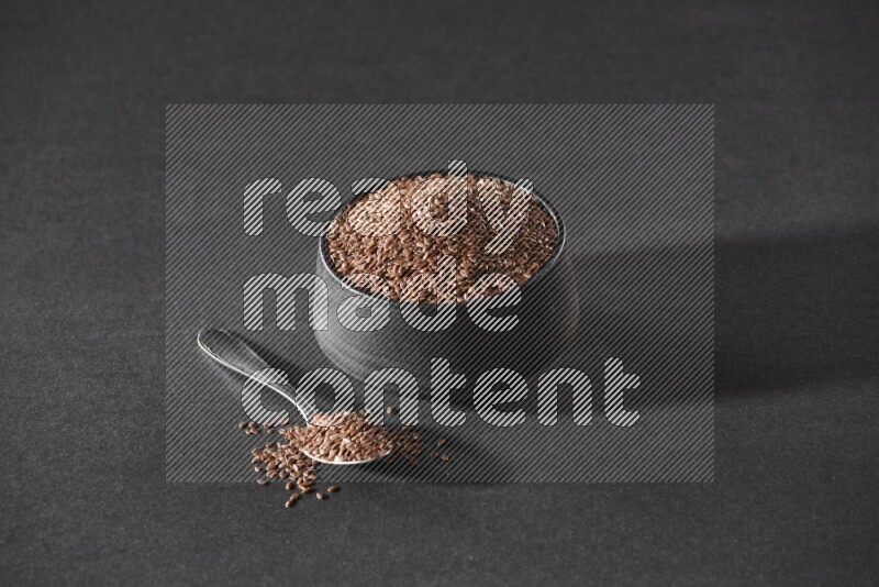 A black pottery bowl full of flaxseeds and a metal spoon full of the seeds on a black flooring