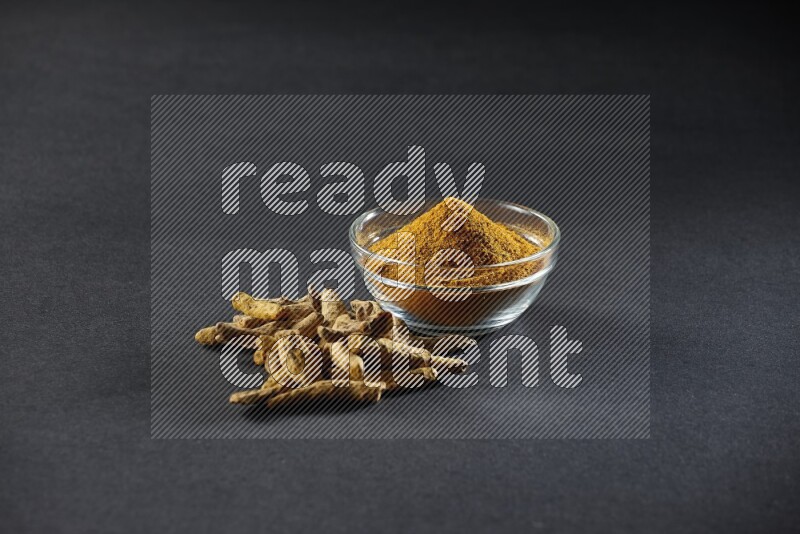 A glass bowl full of turmeric powder and dried whole fingers beside it on black flooring