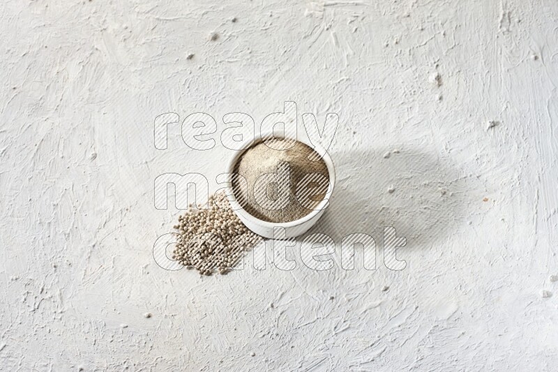 white pottery bowl full of white pepper powder with pepper beads on textured white flooring