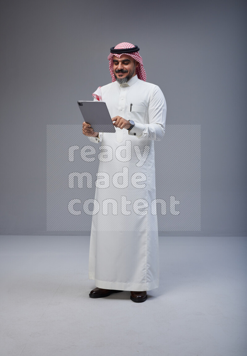Saudi man Wearing Thob and red Shomag standing working on tablet on Gray background