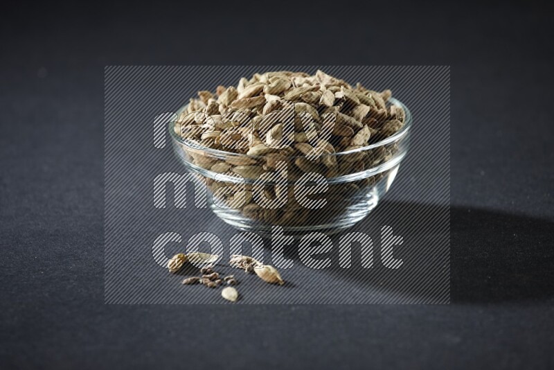A glass bowl full of cardamom and more seeds spreaded beneath the bowl on black flooring