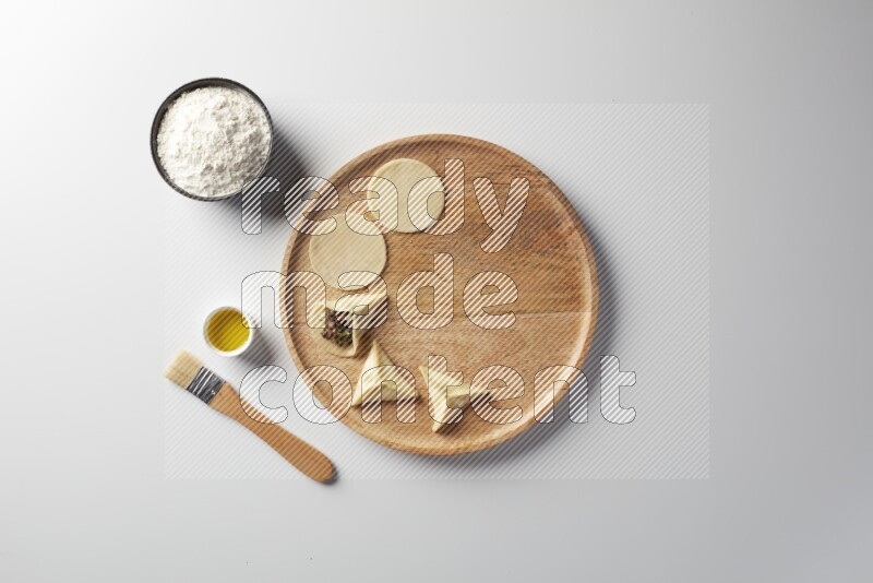 two closed sambosas and one open sambosa filled with meat while flour, and oil with oil brush aside in a wooden dish on a white background