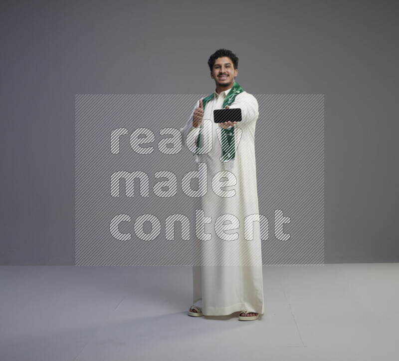A Saudi man standing wearing thob and Saudi flag scarf showing phone to camera on gray background
