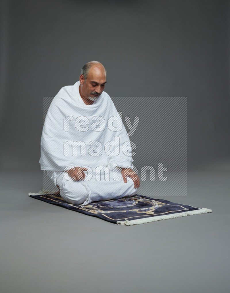 A man wearing Ehram sitting on prayer mat on gray background