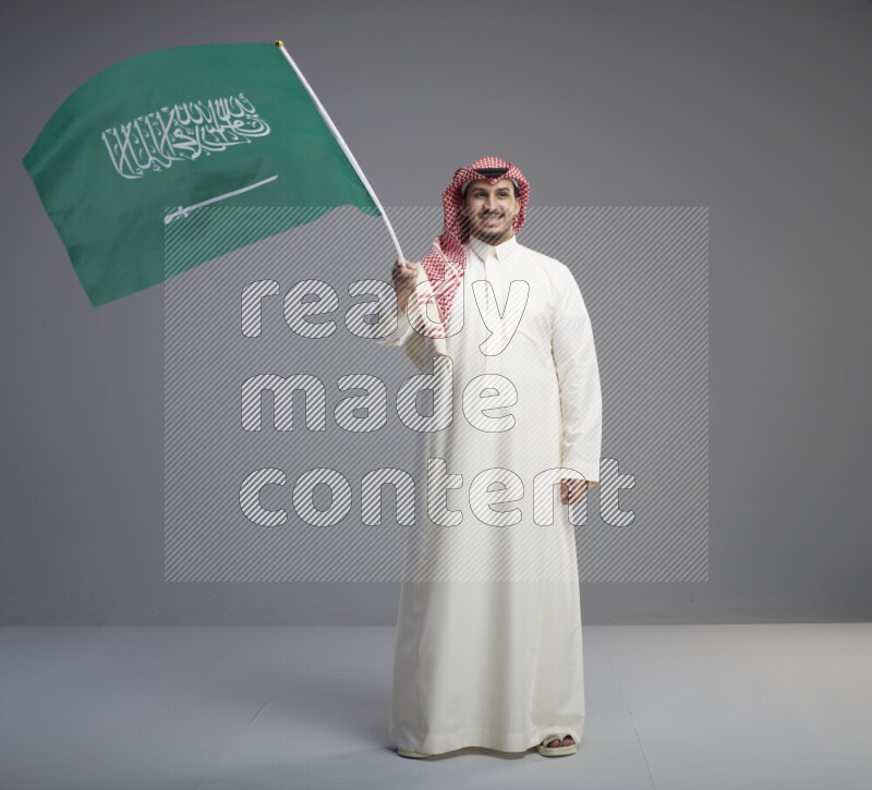 A Saudi man standing wearing thob and red shomag raising big Saudi flag on gray background