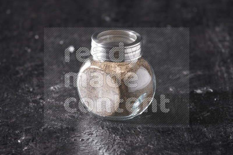 A glass spice jar full of black pepper powder on textured black flooring