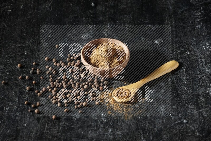A wooden bowl and spoon full of allspice powder and whole balls spreaded on a black flooring