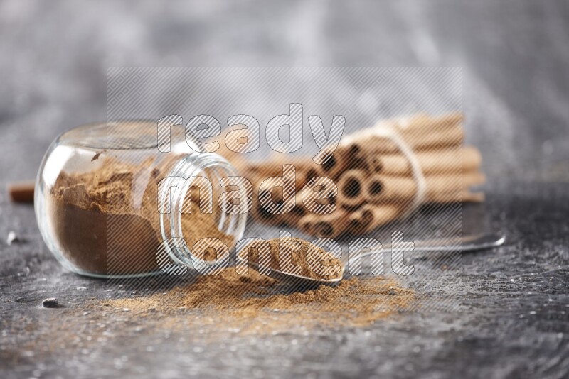 Herbal glass jar full cinnamon powder flipped and a metal spoon full of powder, cinnamon sticks stacked and bounded in the back on textured black background in different angles