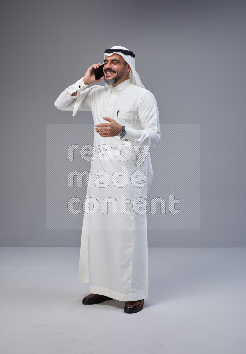 Saudi man Wearing Thob and white Shomag standing talking on phone on Gray background