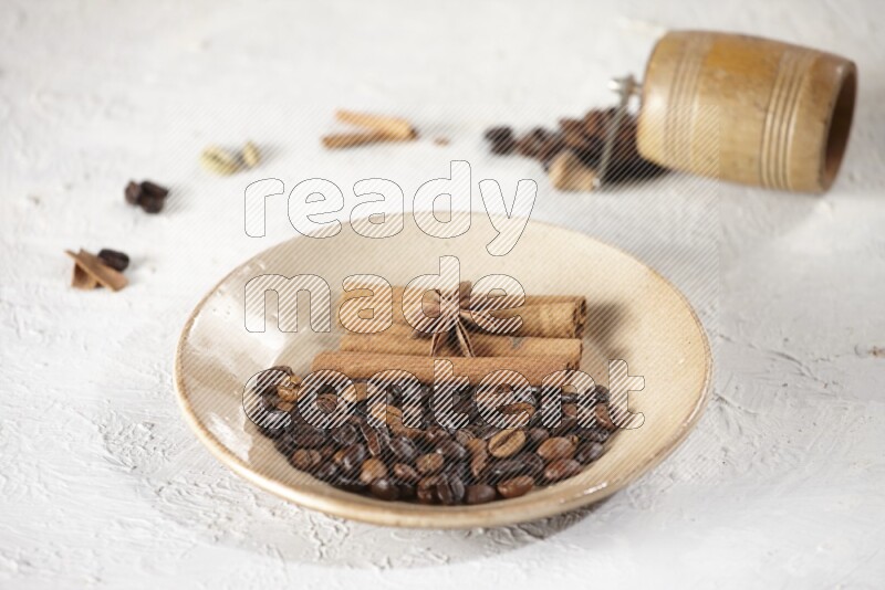 Beige plate full of coffee beans, cinnamon sticks and star anise with a coffee grinder, coffee beans, cinnamon pieces and cardamom next of it on white background