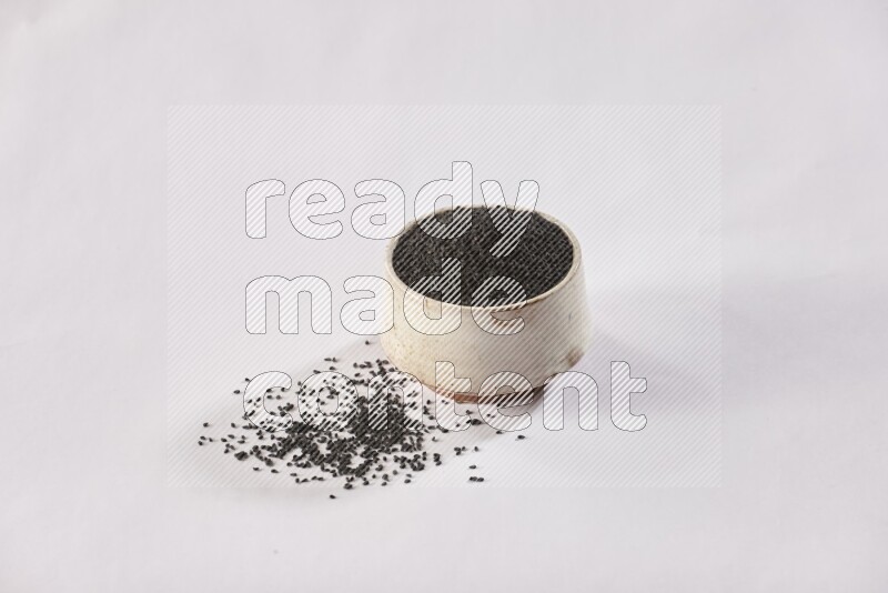 وعاء بيج فخاري ممتلئ ببذور حبة البركة مع المزيد من البذور المتناثرة علي خلفية بيضاء