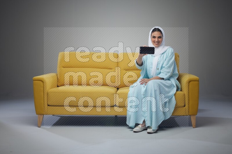 A Saudi woman wearing a light blue Abaya and white head scarf sitting on a yellow sofa and showing her phone's screen vertically and horizontally eye level on a grey background