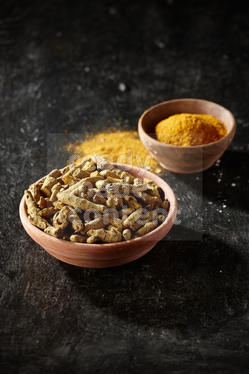 2 wooden bowls, one full of turmeric powder and the other full of dried turmeric whole fingers on a textured black flooring