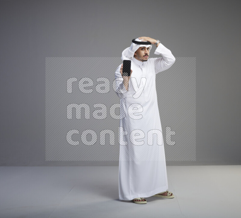 A Saudi man standing wearing thob and white shomag showing phone to camera on gray background