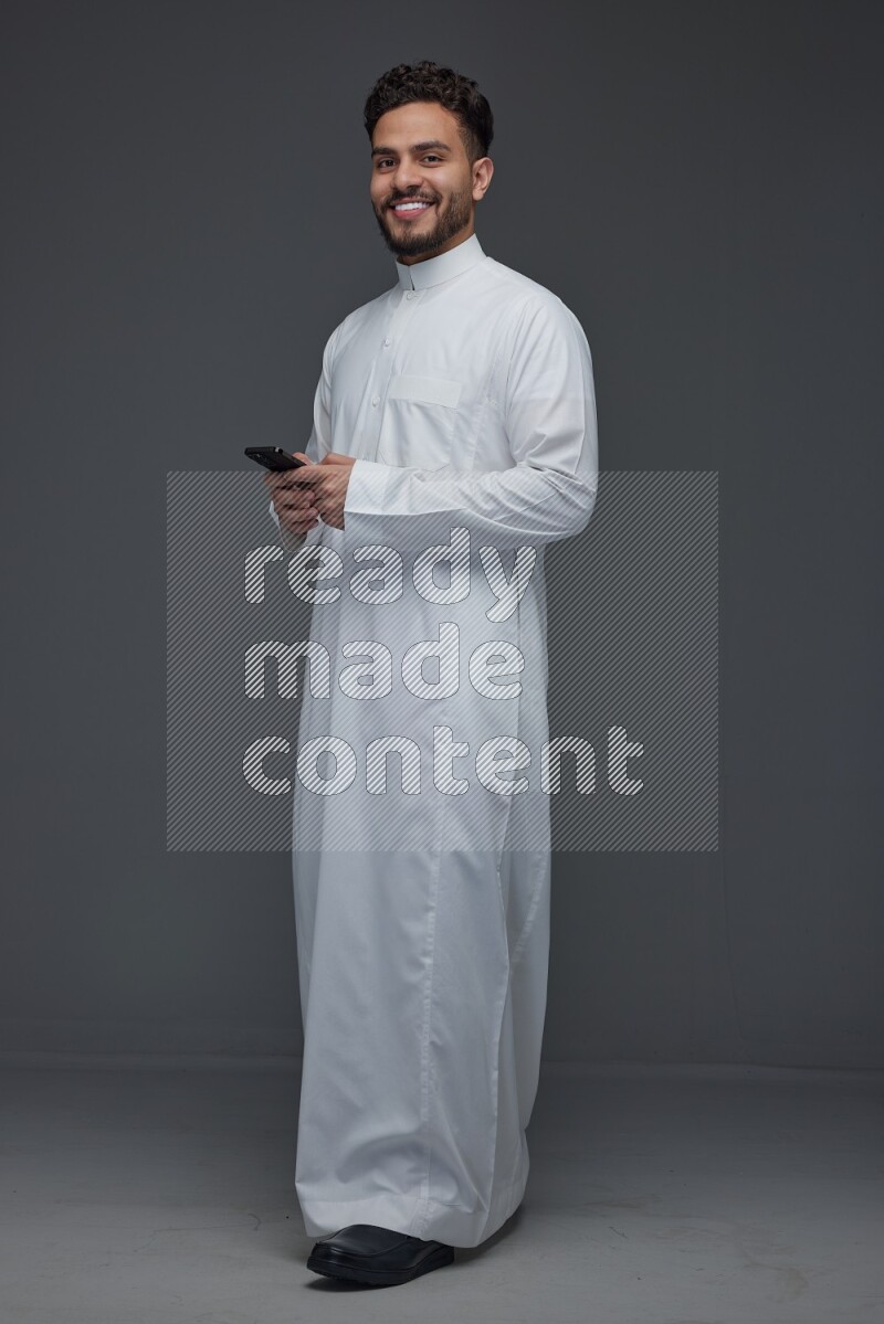 A Saudi man wearing Thobe standing and using his phone eye level on a gray background
